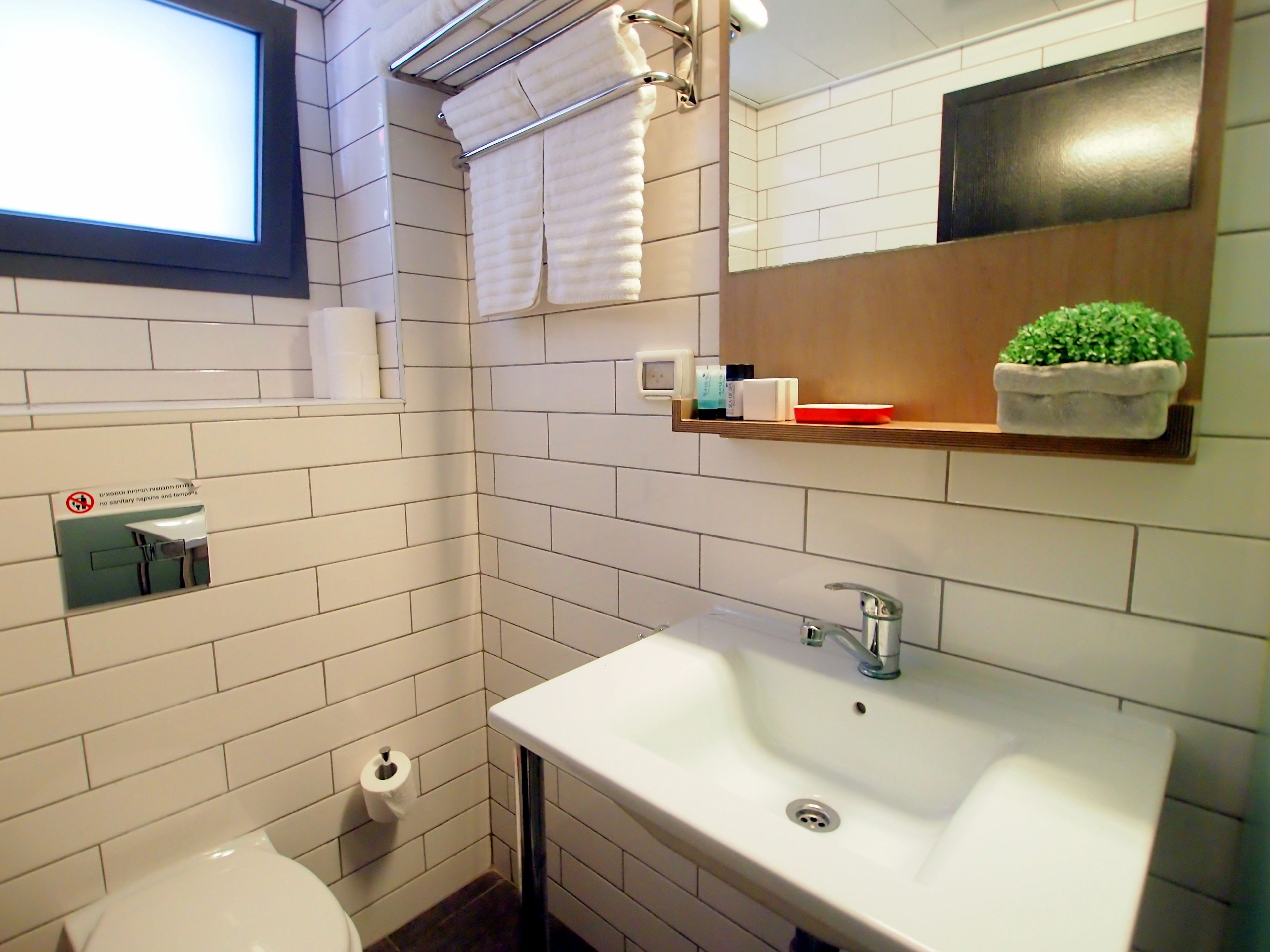 A bathroom with white tiles, a sink, a mirror, a towel rack, a toilet, and a small shelf with toiletries.