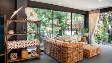 Un salón moderno con un sofá de mimbre, un estante de madera con objetos decorativos y vistas a un exuberante follaje exterior.