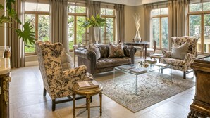 A spacious living room with a leather sofa, floral armchairs, a glass coffee table, and a large window with curtains.