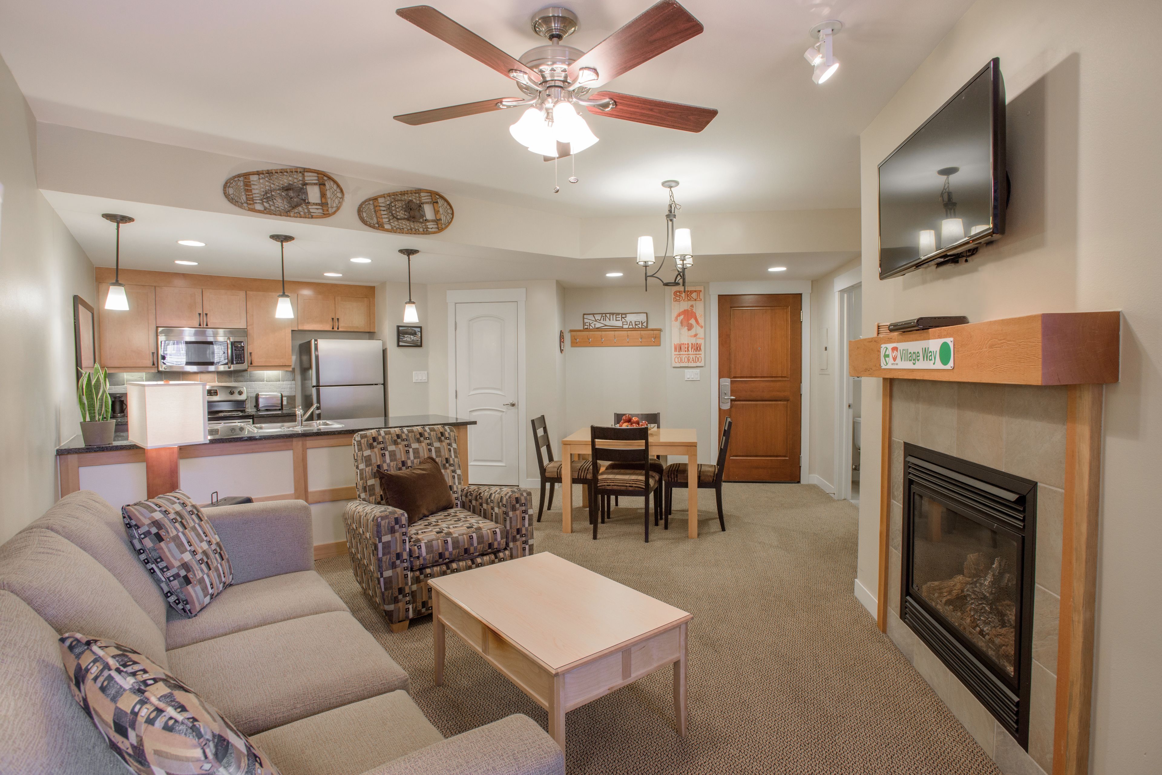 A modern living room with a sofa, a coffee table, a fireplace, a TV mounted on the wall, a dining area with chairs, and a kitchen area with appliances.