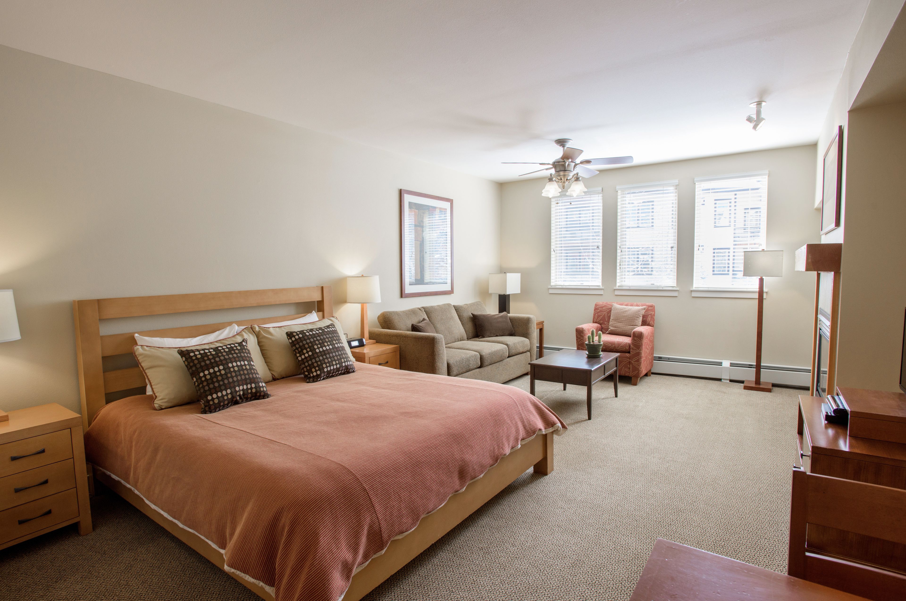 A bedroom with a large bed, a sofa, a chair, and a desk. There are windows with blinds, a ceiling fan, and a floor lamp.