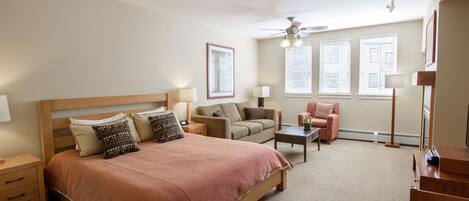 A bedroom with a large bed, a sofa, a chair, and a desk. There are windows with blinds, a ceiling fan, and a floor lamp.