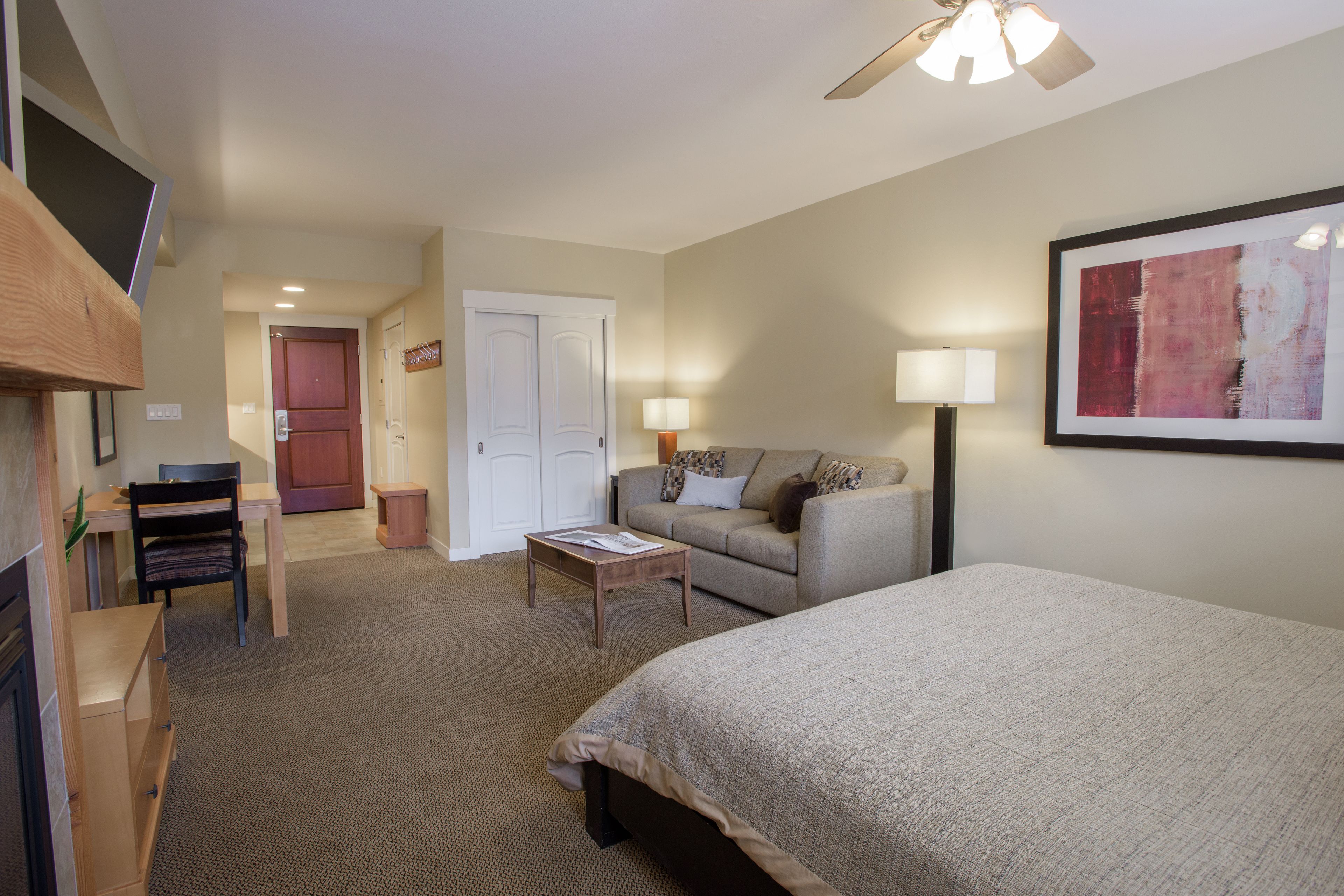 A hotel room with a bed, a sofa, a desk with a chair, a small table, a lamp, and a ceiling fan.