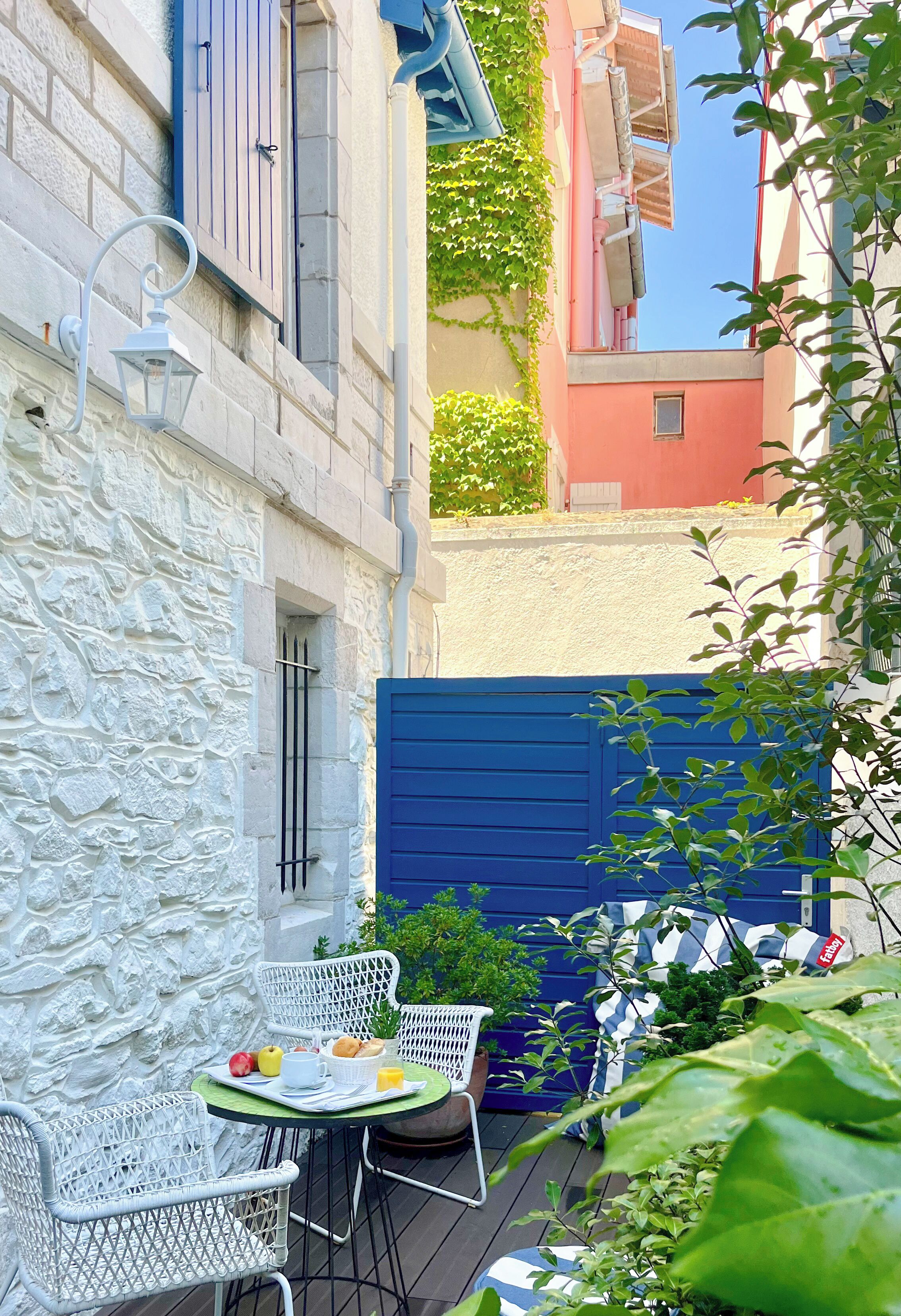 A narrow outdoor patio with a table set for breakfast, surrounded by white metal chairs and a blue gate.