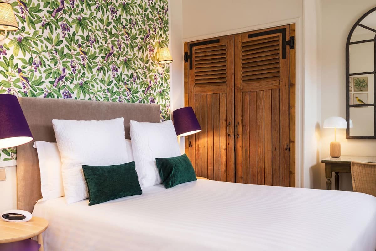 A hotel room with a bed, two lamps, a wooden door, and a wall with a floral pattern.