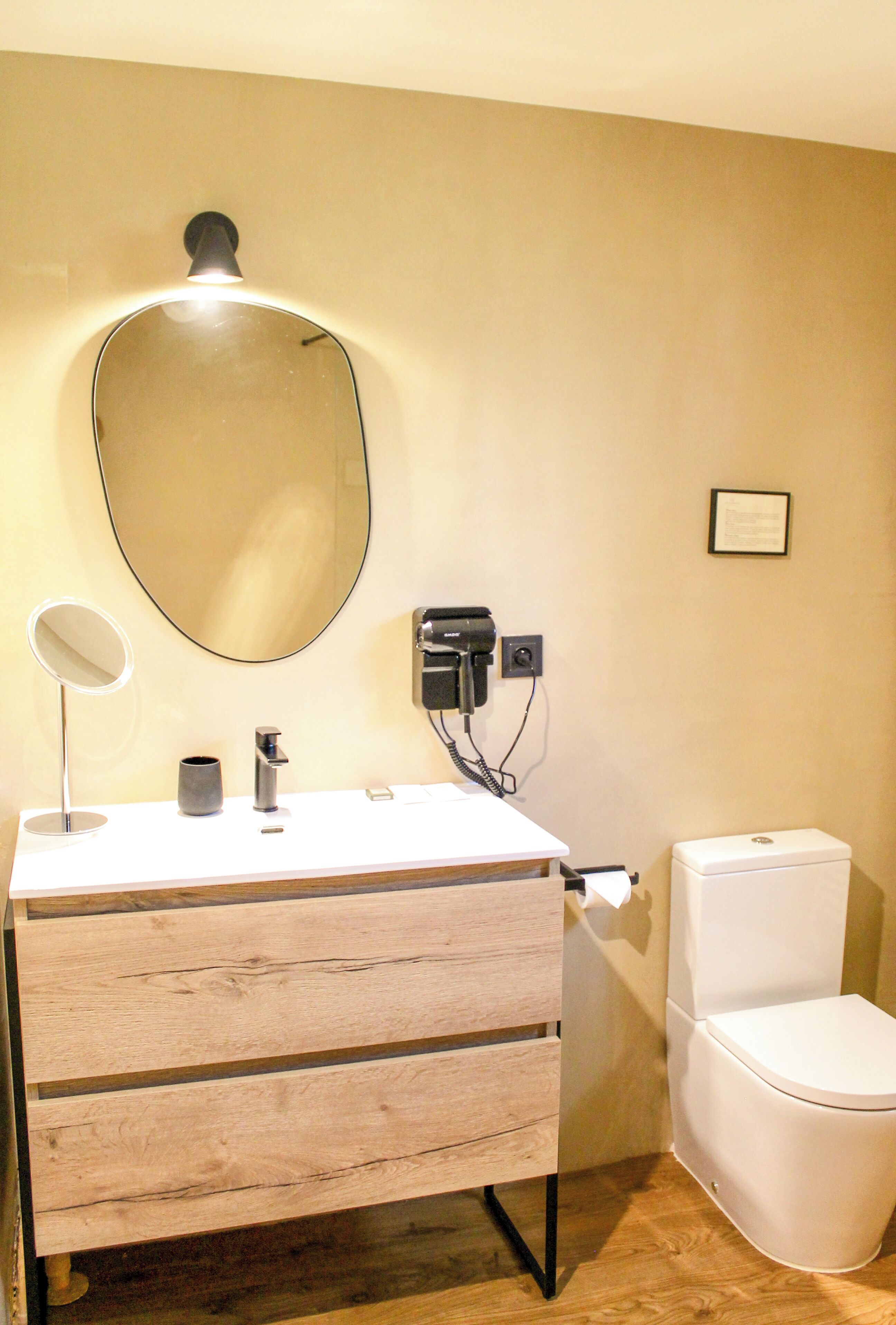 Un baño con tocador de madera, espejo redondo, secador de pelo y inodoro.