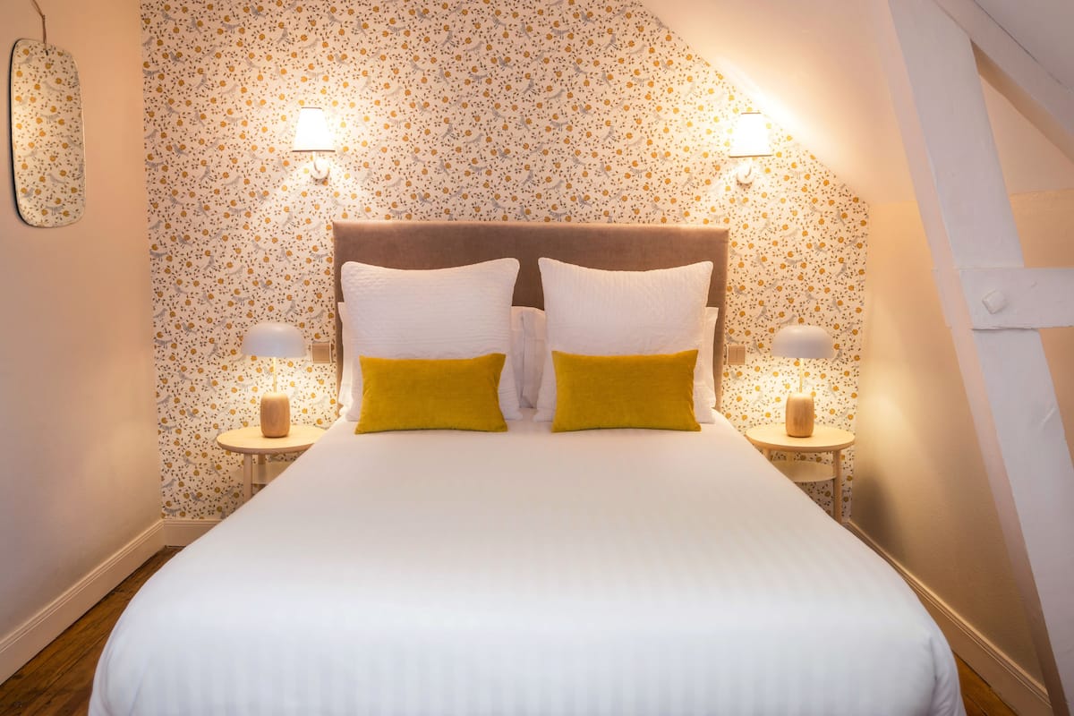 A neatly made bed with white and yellow pillows, two bedside lamps, and floral wallpaper.