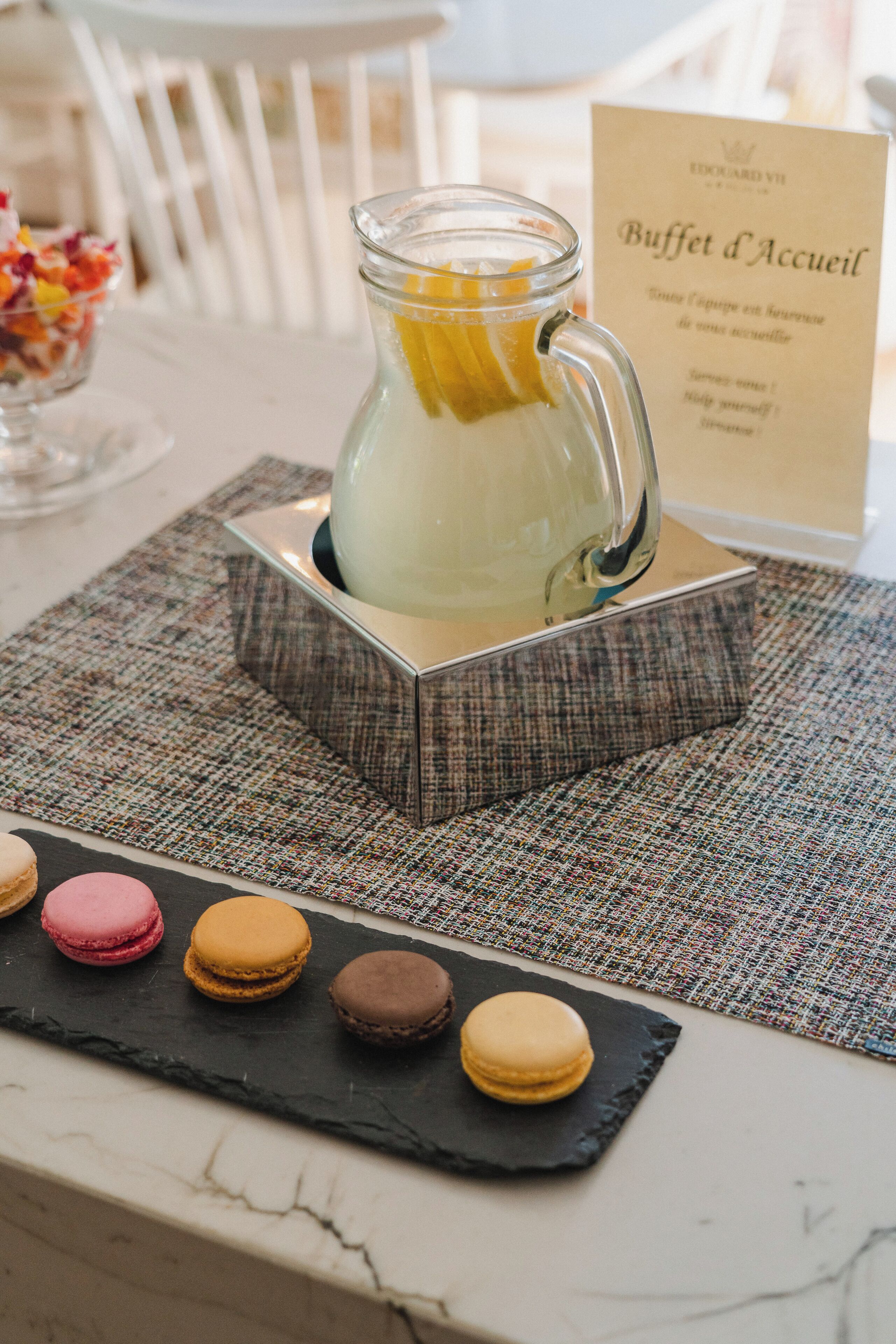 A table with a glass pitcher of lemonade, a sign reading "Buffet d'Accueil," and macarons on a slate board.