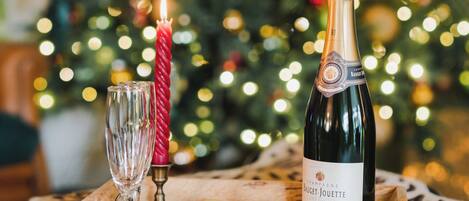 A table with a bottle of champagne, a glass, a candle, and cookies in front of a decorated Christmas tree.