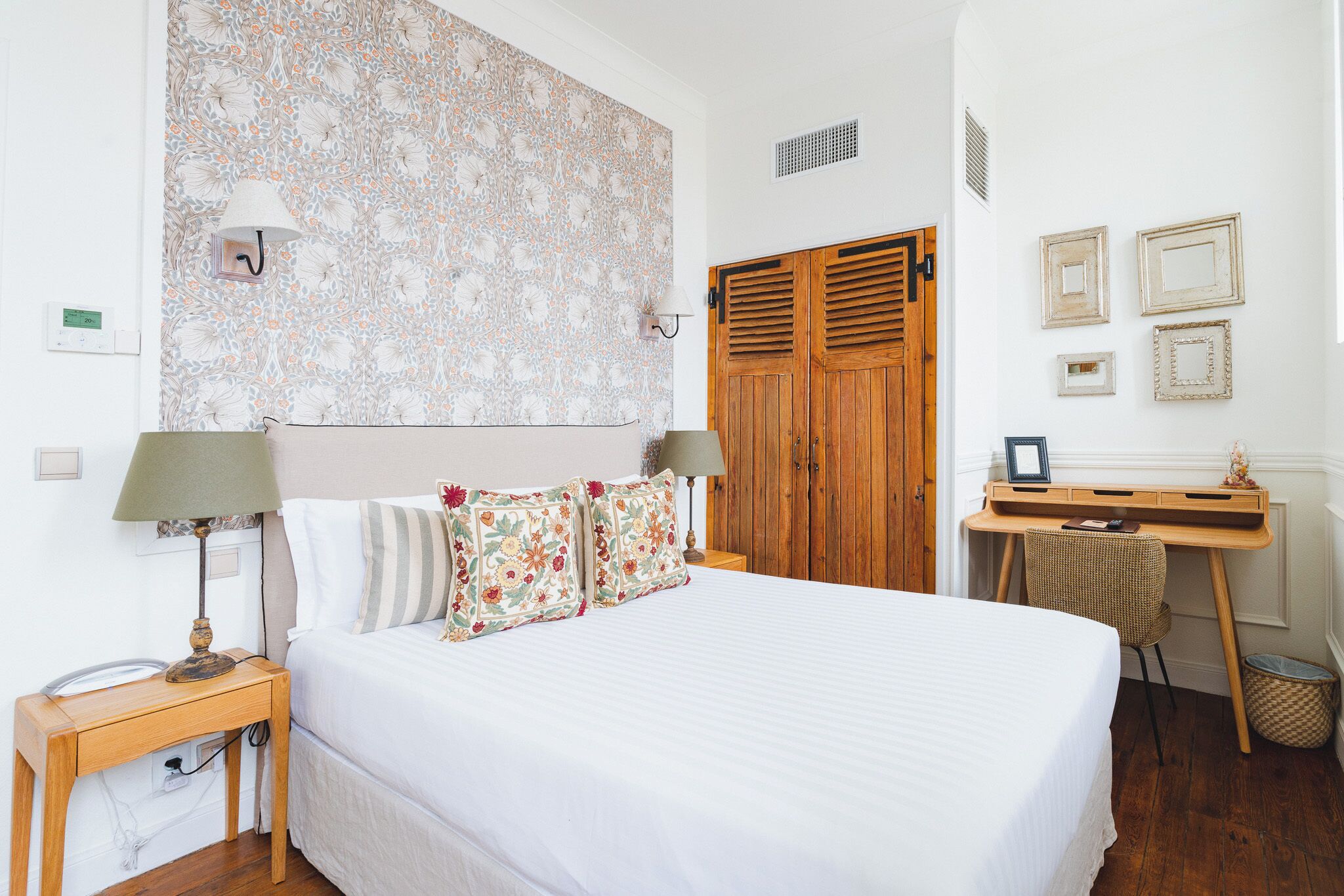 A bedroom with a bed, a wooden desk, a wardrobe, and a patterned wall.
