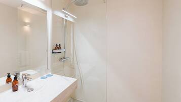 Un baño moderno con cabina de ducha de vidrio, lavamanos blanco con espejo y estante con artículos de aseo.
