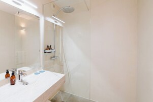 Un baño moderno con cabina de ducha de vidrio, lavamanos blanco con espejo y estante con artículos de aseo.