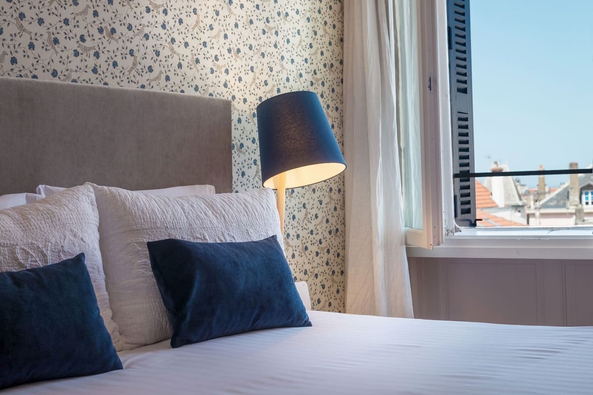 A neatly made bed with pillows, a lamp, and a window with a view of buildings.