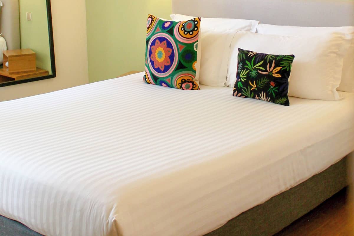 A neatly made bed with white linens and two decorative pillows, a bedside table with a lamp, and a mirror on the wall.