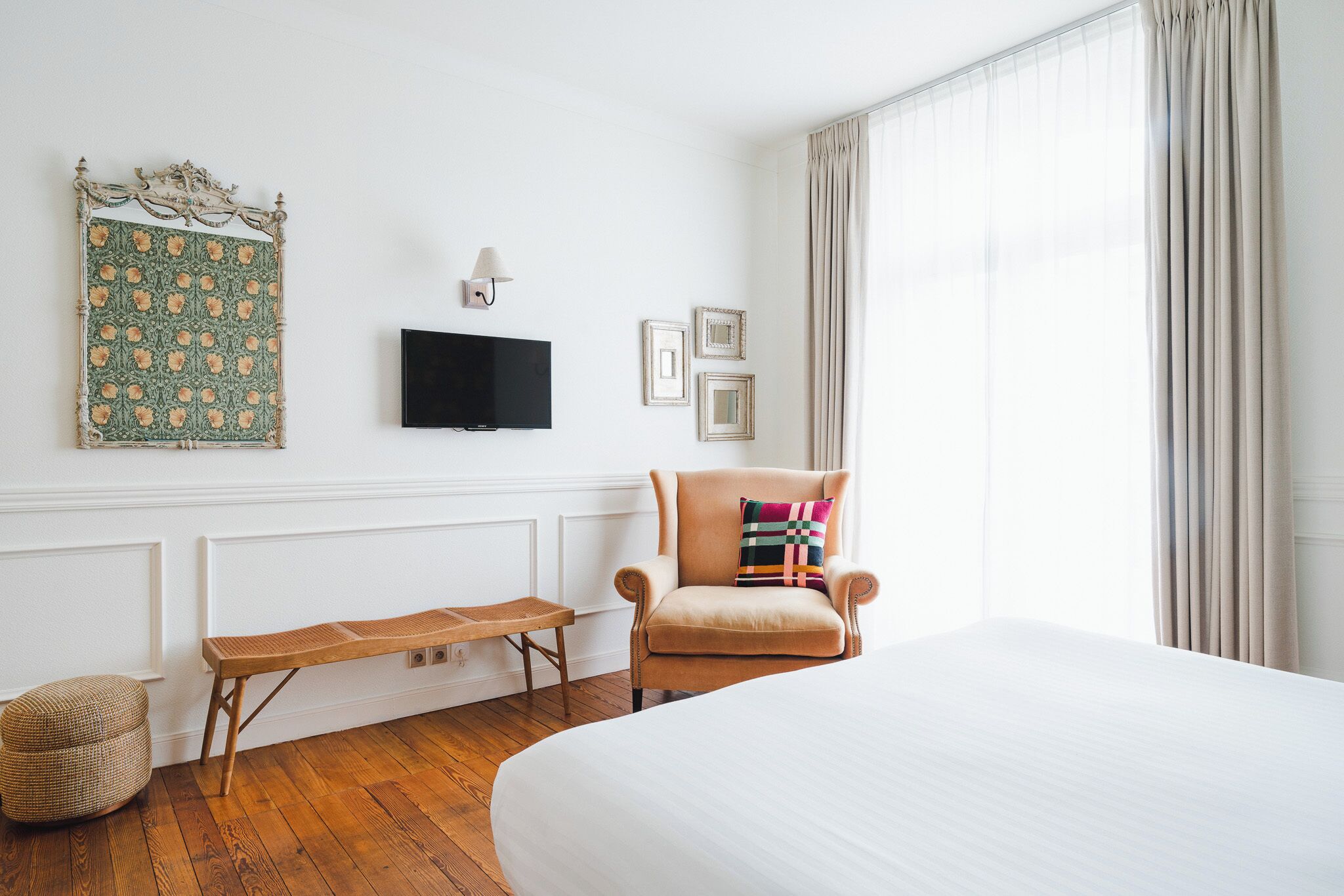 Une chambre d’hôtel avec un grand lit, un banc en bois, une chaise avec un coussin coloré, un téléviseur fixé au mur et une tapisserie décorative.
