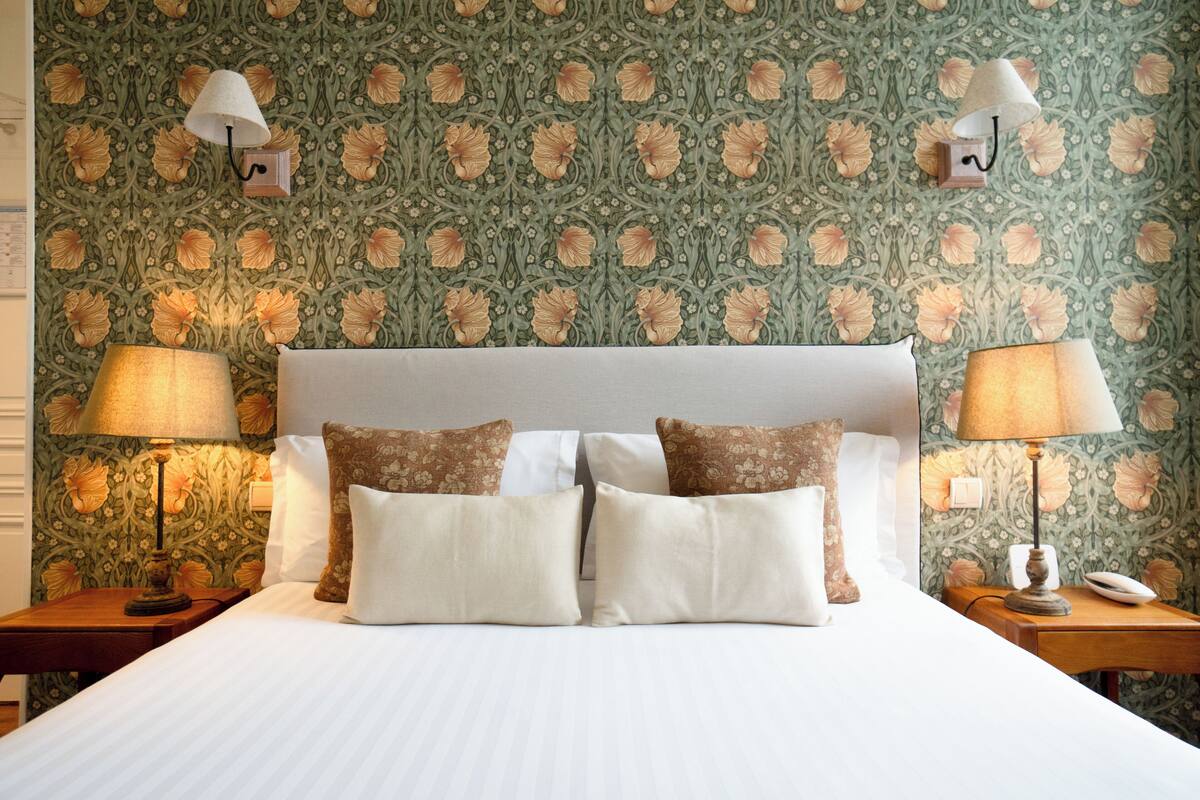 A neatly made bed with white linens and two decorative pillows, flanked by two bedside tables with lamps, set against a wallpapered wall with a floral pattern.