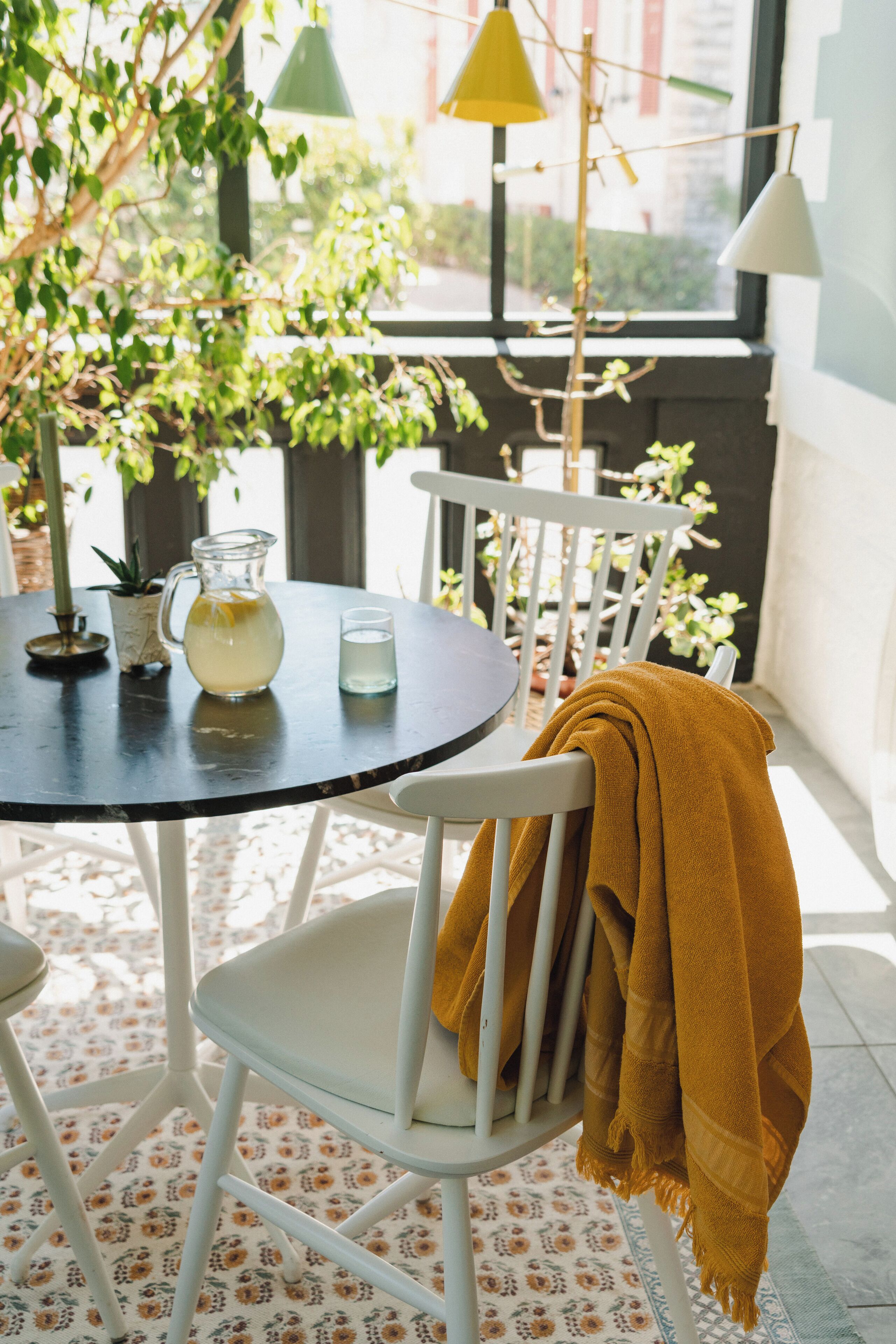 Une table ronde avec une carafe et un verre, une chaise blanche recouverte d’un plaid jaune et un sol à motifs.
