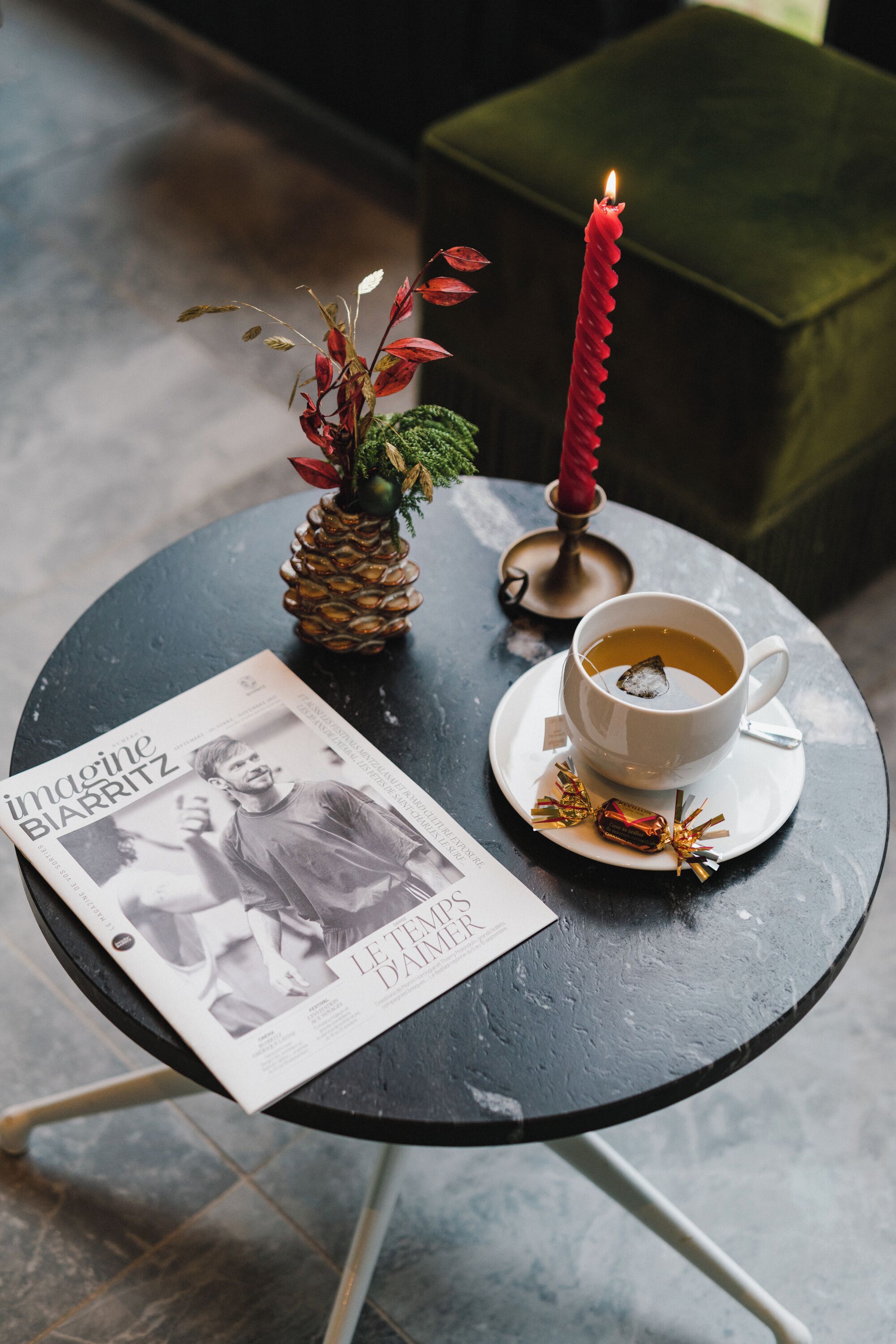 A table with a cup of tea, a red candle, a pineapple, and a magazine.