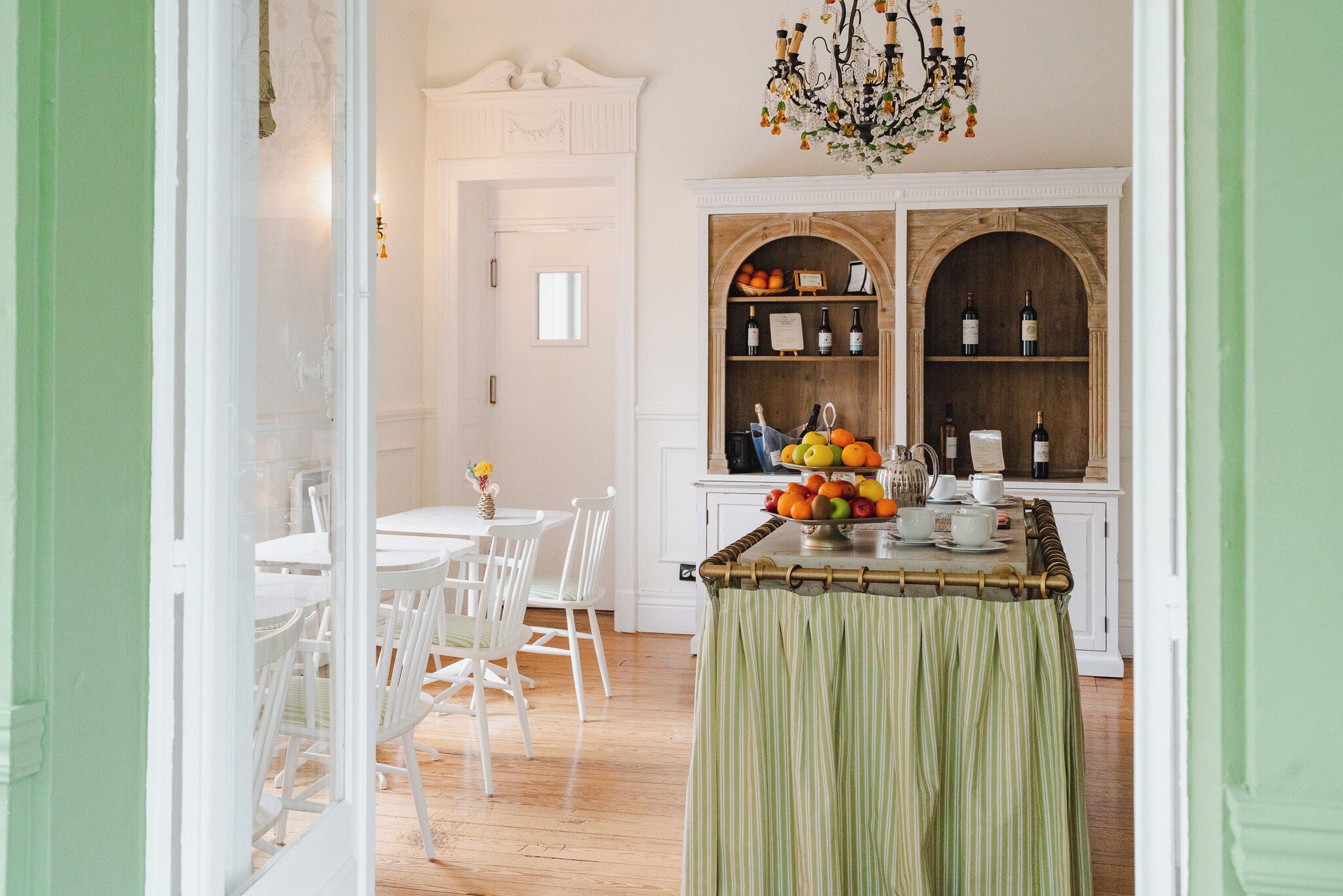 A dining area with a wooden table, chairs, and a fruit bowl. There are two wooden shelves with wine bottles and a chandelier above.