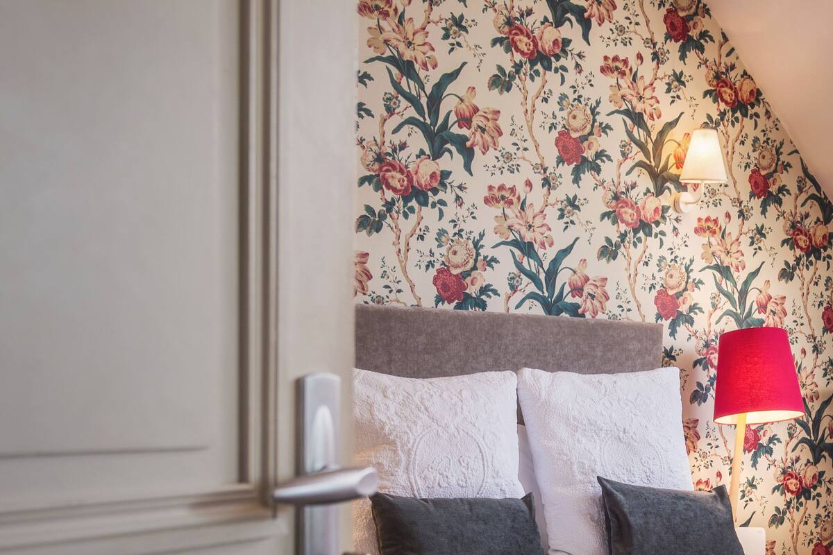A bedroom with a floral wallpaper, a bed with white and dark grey bedding, a red lamp, and a door with a lock.