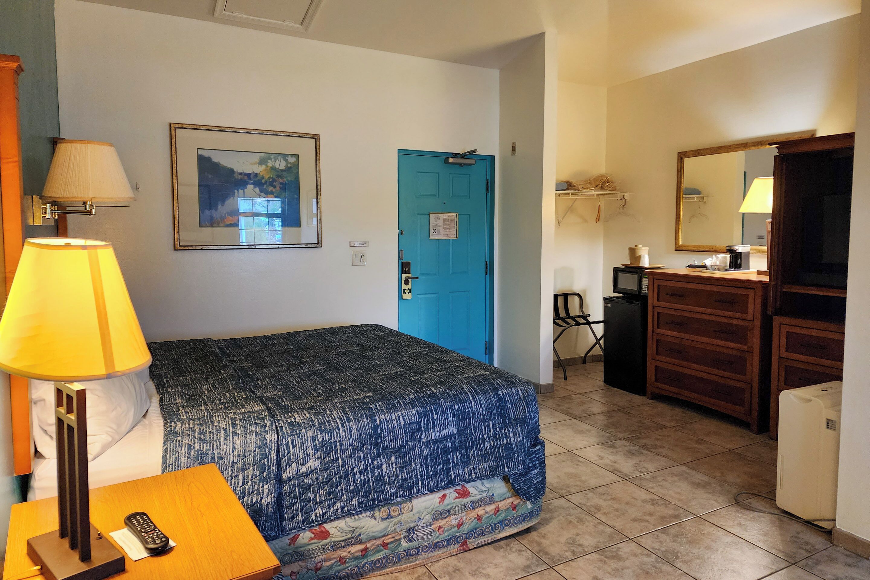 A hotel room with a bed, a nightstand with a lamp, a dresser, a mirror, and a door.