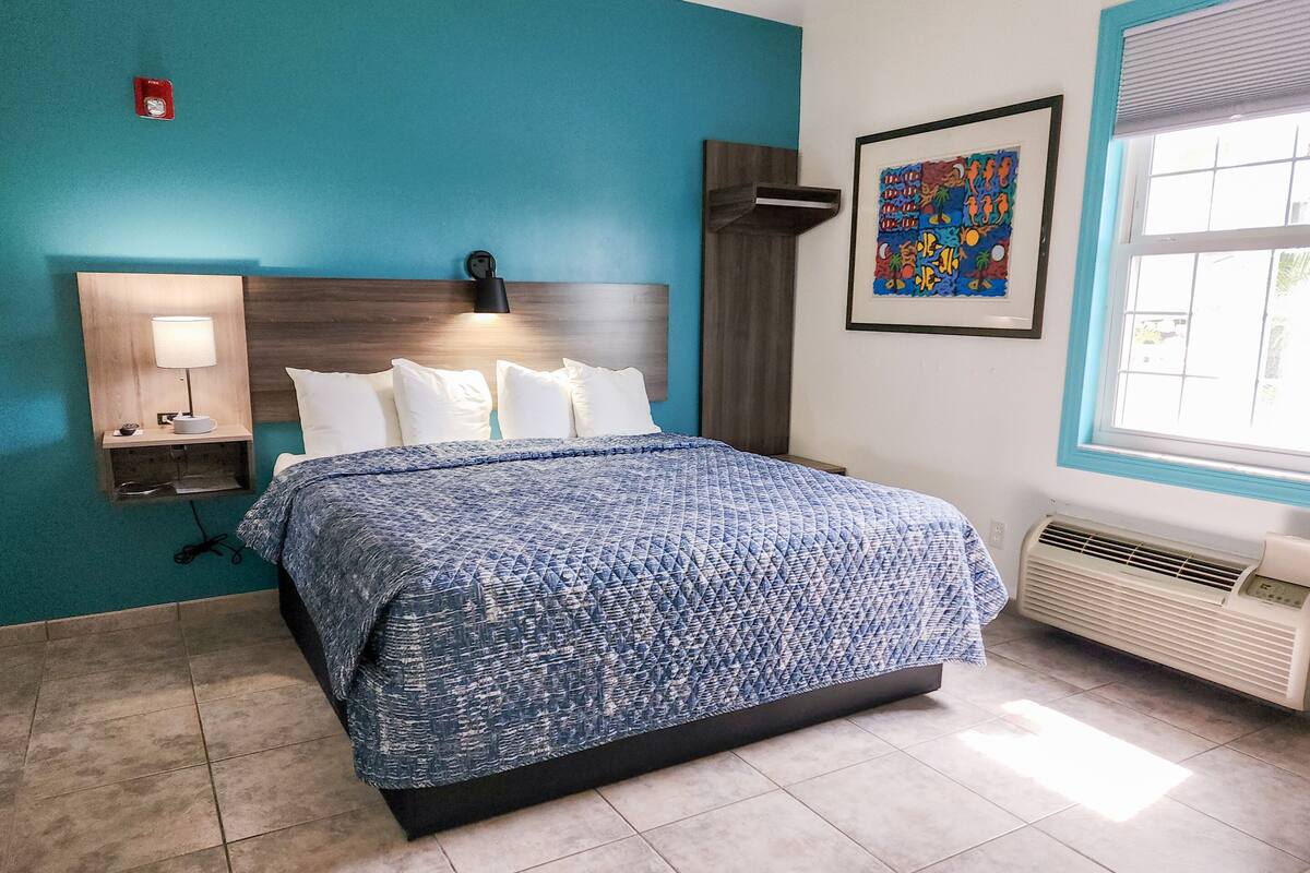 A hotel room with a bed, a nightstand, a wall-mounted lamp, a framed artwork, and a window with blinds.