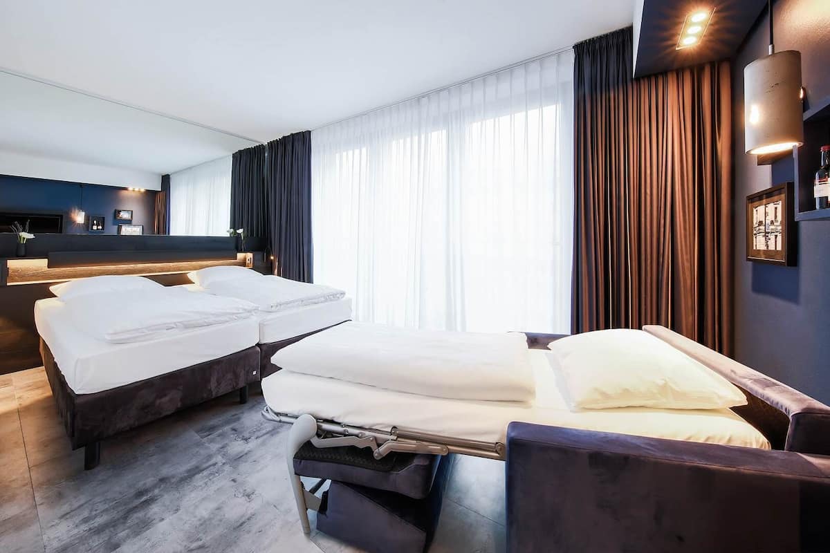 Une chambre d’hôtel avec deux lits, une grande fenêtre avec des rideaux, une table de chevet et une lampe fixée au mur.
