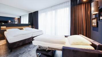Quarto de hotel com duas camas, uma janela grande com cortinas, uma mesinha de cabeceira e uma luminária fixada na parede.