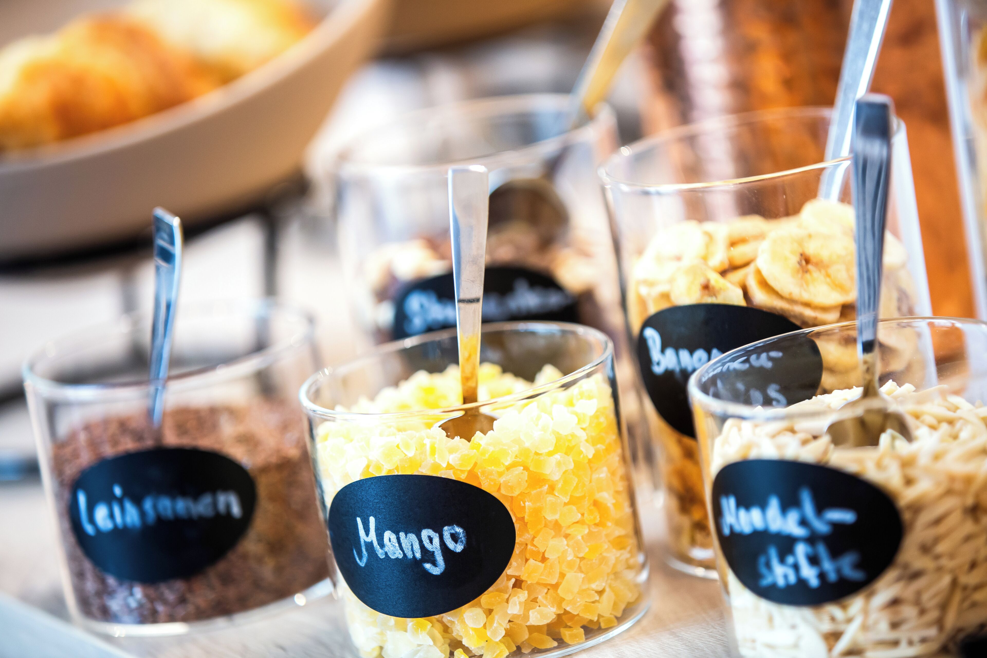 Uma seleção de alimentos em copos transparentes, com etiquetas indicando 'leinsamen', 'Mango' e 'Hoodel-shifte'.