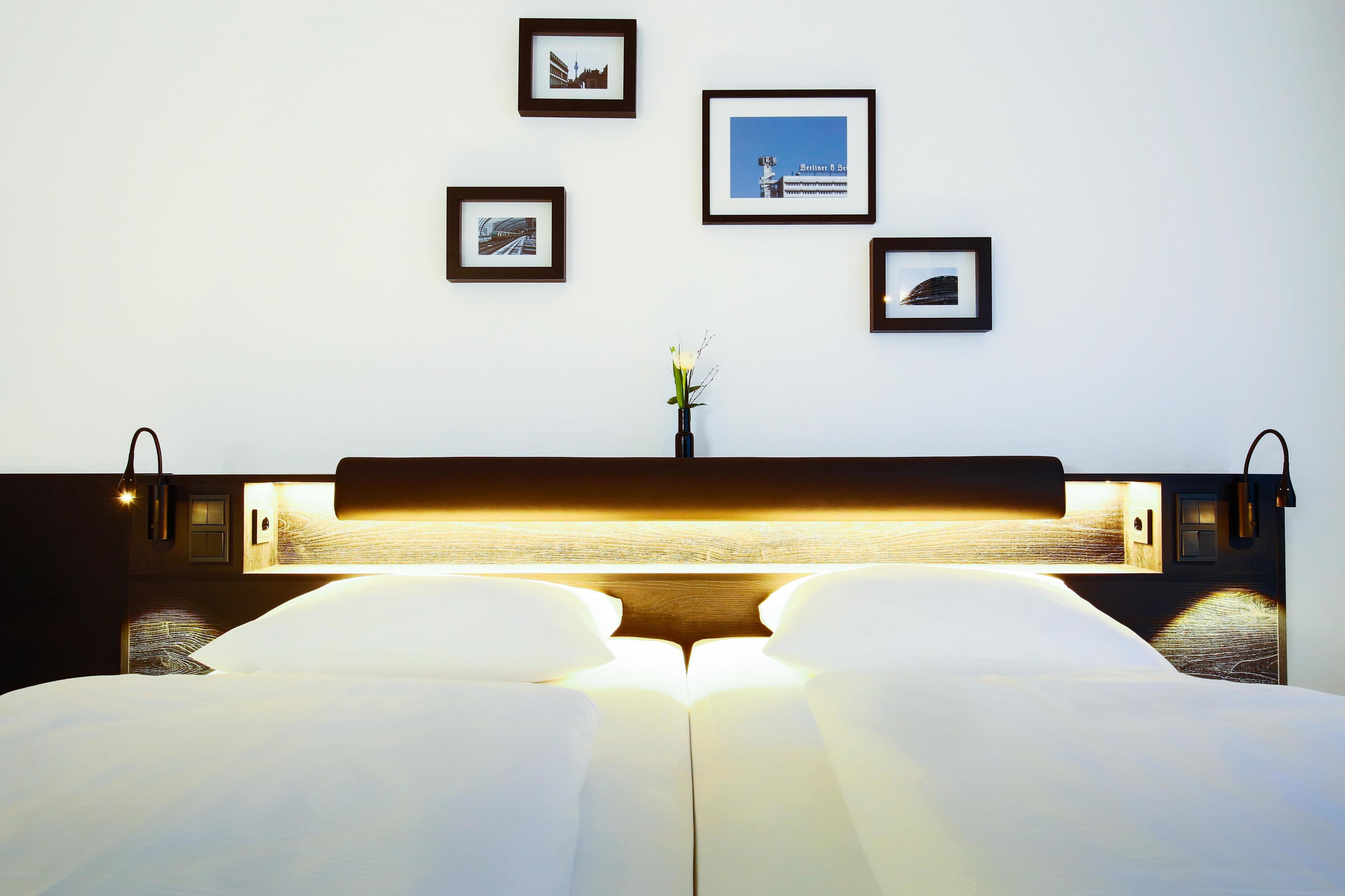 Una cama bien hecha con sábanas blancas, una mesita de noche con una lámpara y cuadros en la pared.
