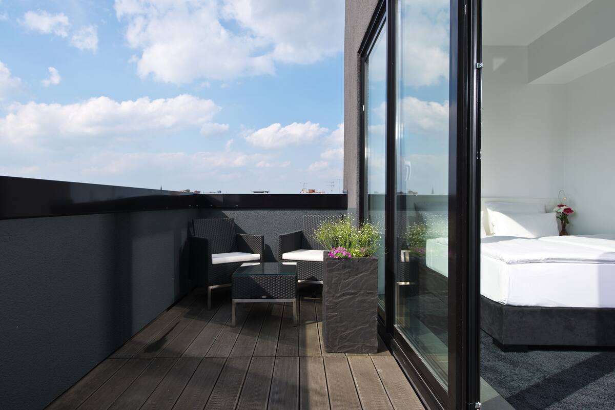 Un balcon avec une porte-fenêtre donnant sur une chambre, cette dernière étant équipée d’un lit, d’une petite table et de deux chaises.