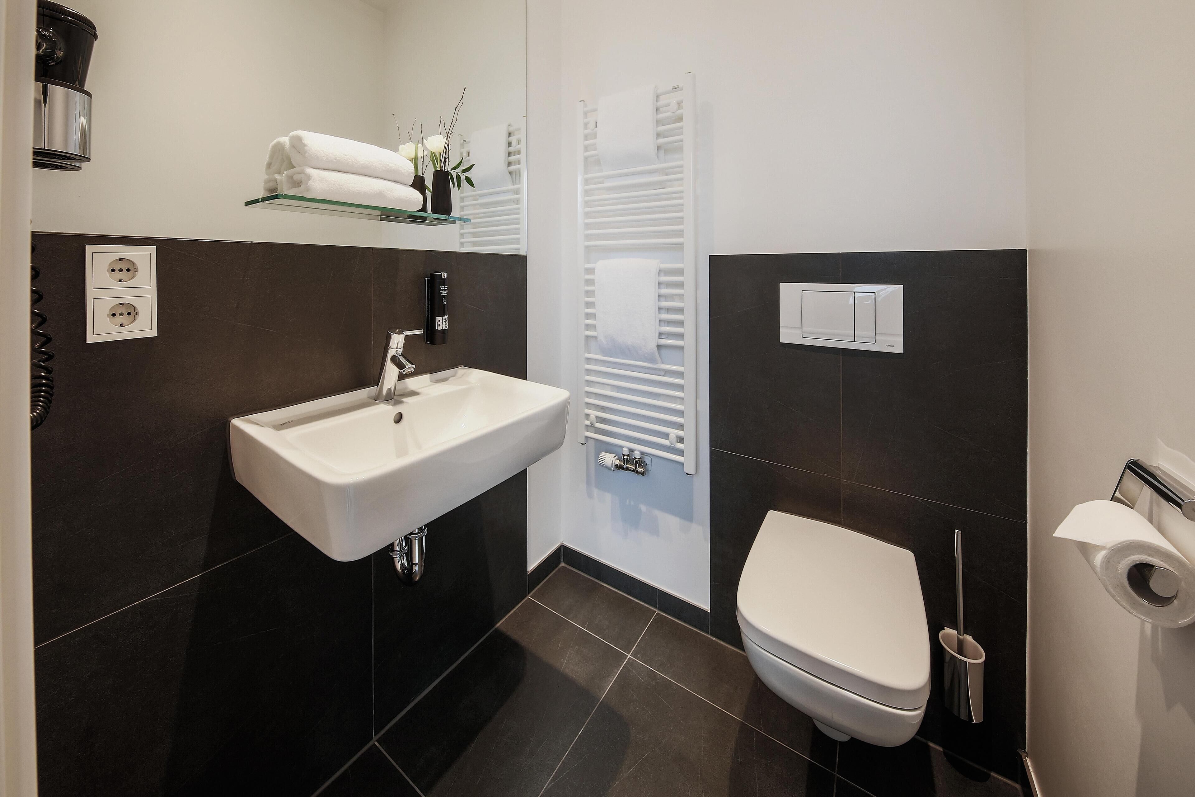 Une salle de bain moderne avec un lavabo blanc, des murs recouverts de carreaux foncés, des toilettes blanches et un porte-serviettes.