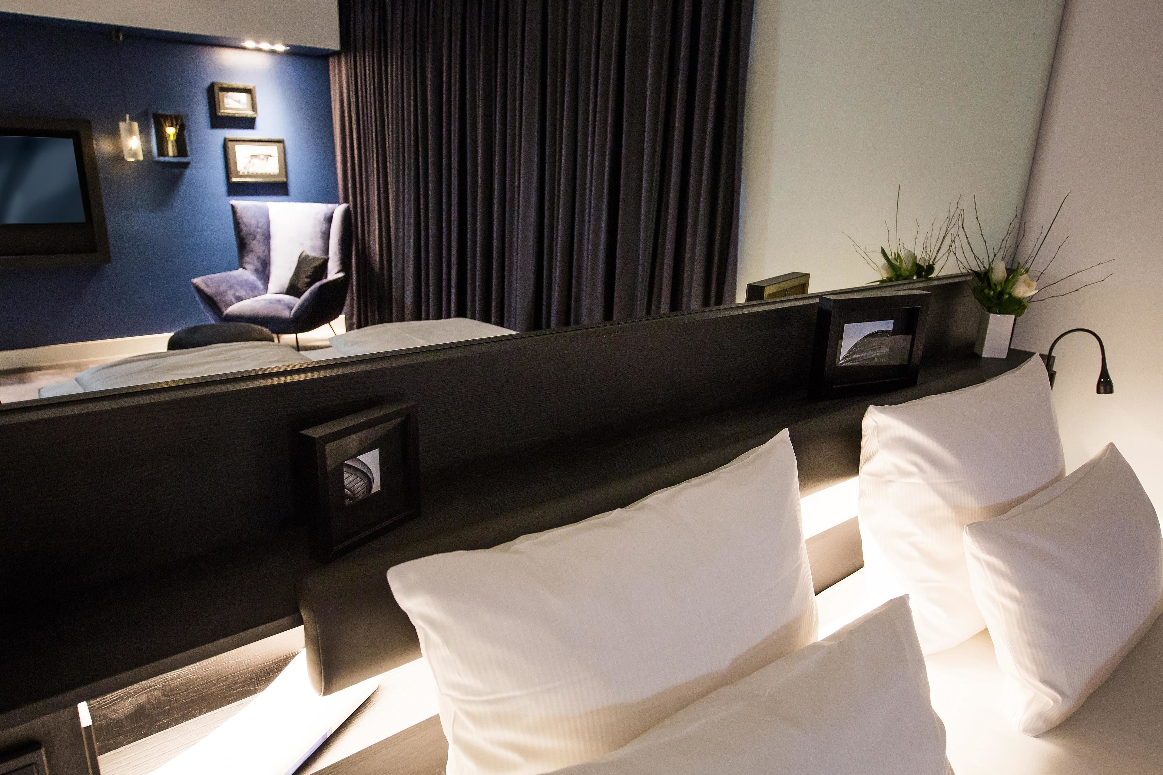 A modern hotel room with a dark blue accent wall, a large flat-screen TV, a comfortable armchair, a bedside table with a lamp, and a large bed with white linens.