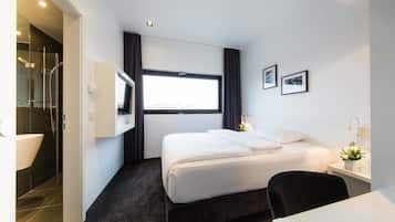 A modern hotel room with a large bed, a desk, a bathroom with a glass shower, and a window providing natural light.