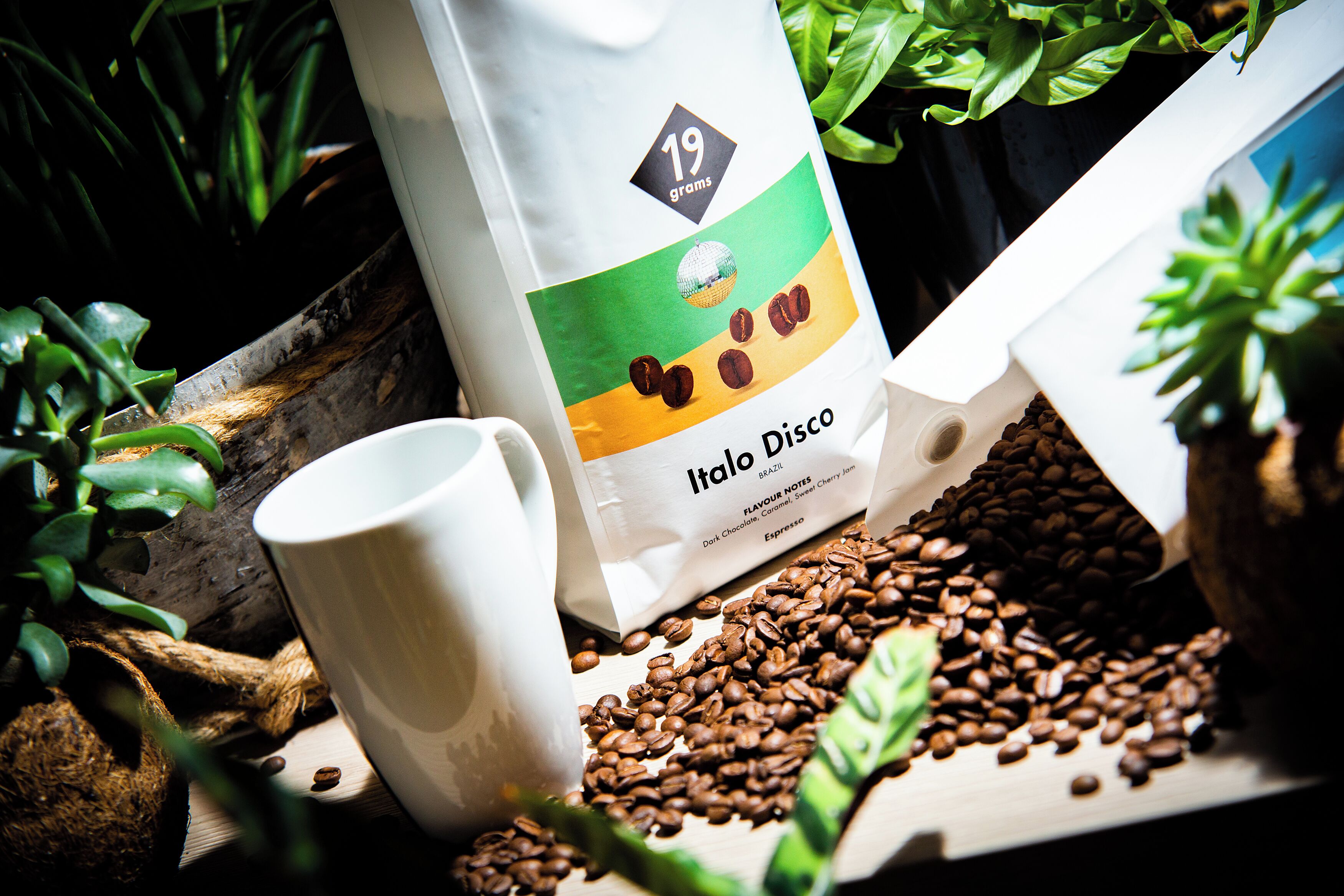 A coffee bag labeled 'Italo Disco' with a 19-gram weight indication, a white mug, and scattered coffee beans.