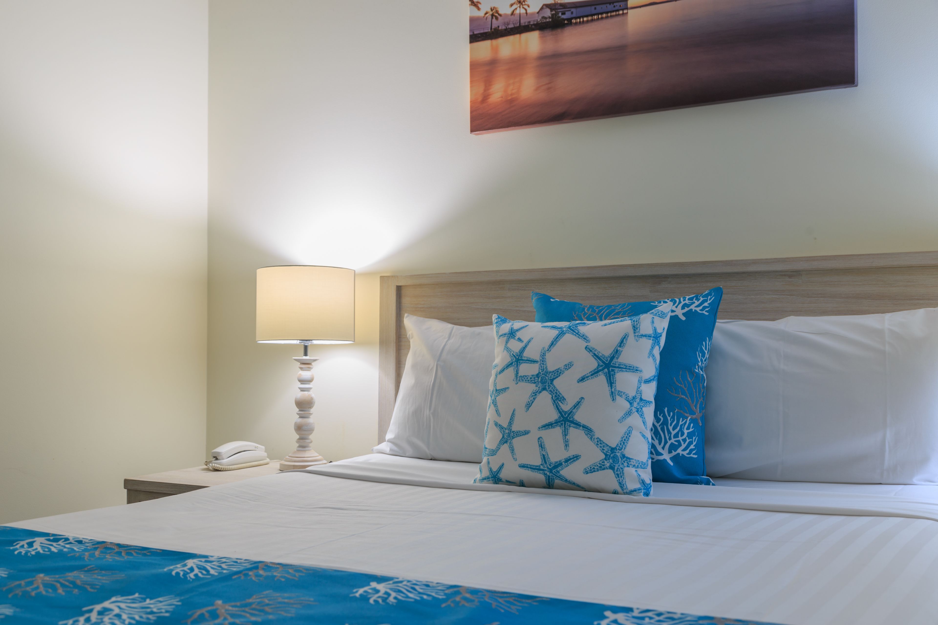 A neatly made bed with white linens and blue patterned pillows, a bedside lamp, and a painting on the wall.