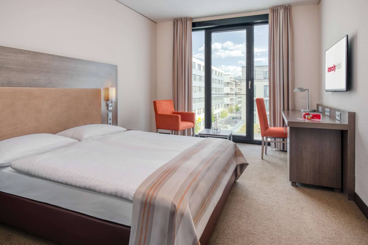 Ein Hotelzimmer mit einem großen Bett, einem Schreibtisch, zwei Stühlen, einem Fernseher und Blick auf Gebäude durch das Fenster.