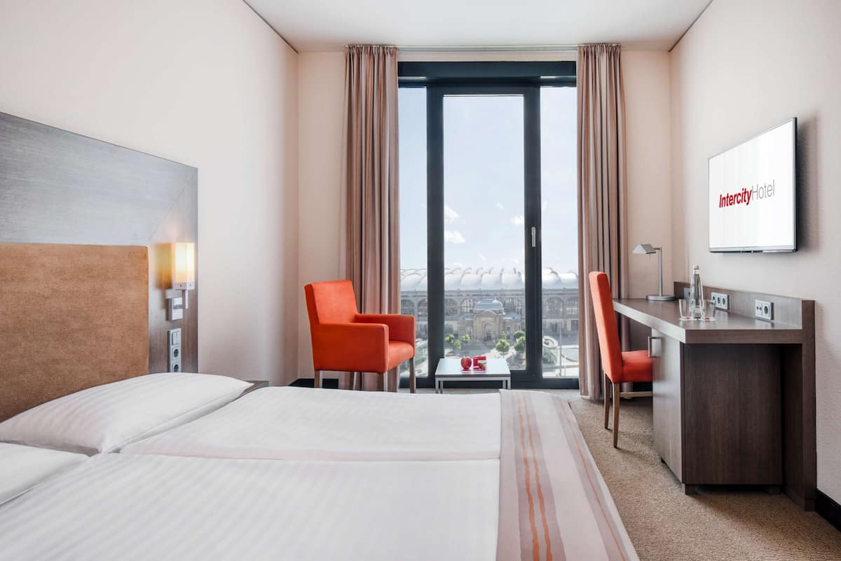 Ein Hotelzimmer mit einem Bett, zwei Stühlen, einem Schreibtisch, einem Fernseher und Blick auf eine Stadtansicht.