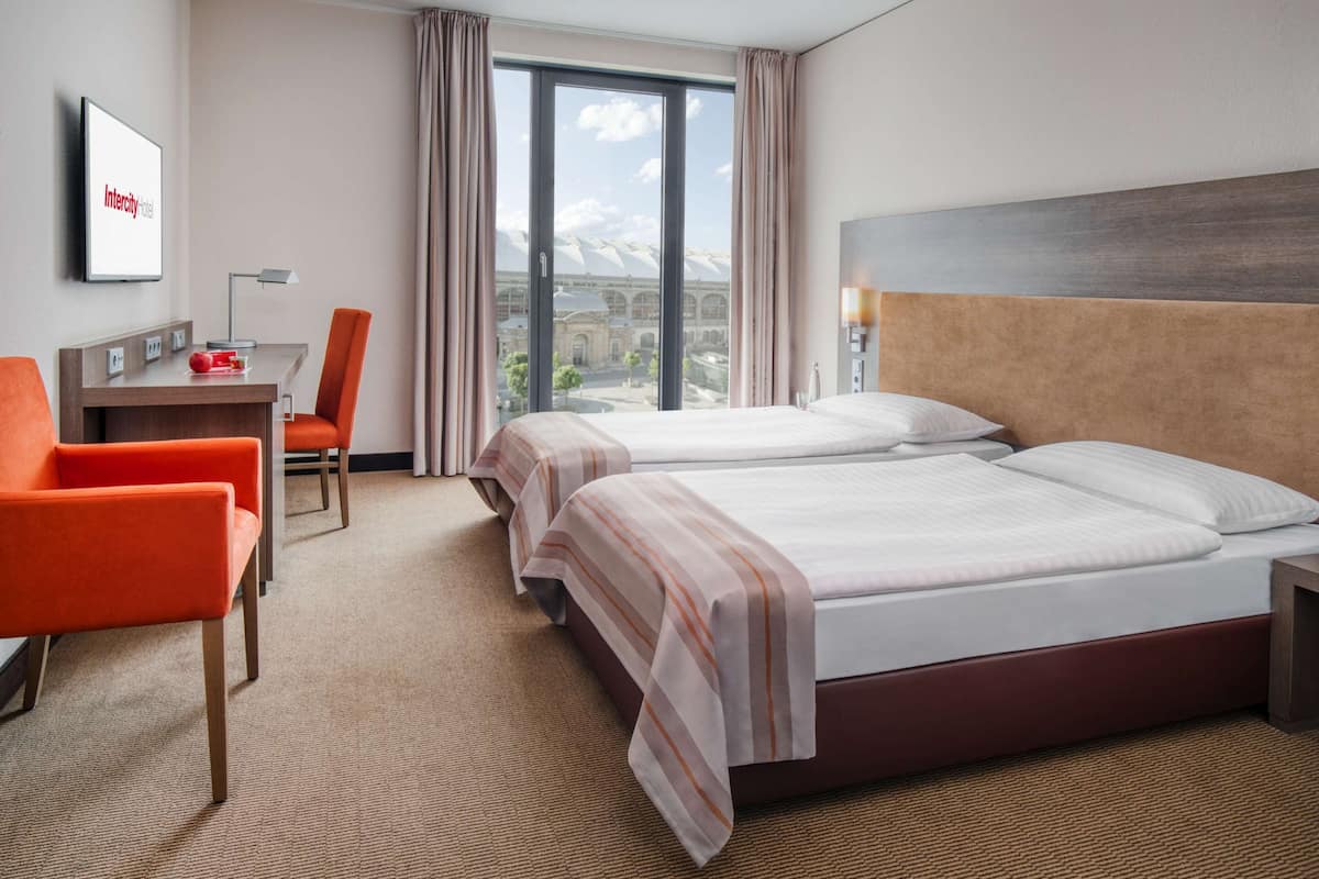 Ein Hotelzimmer mit zwei Betten, einem Schreibtisch mit Stuhl, einem Fernseher und einem großen Fenster mit Blick auf ein Gebäude.