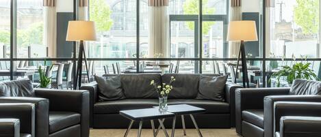 A modern hotel lobby with leather sofas, a central coffee table, and large windows offering a view of the city.
