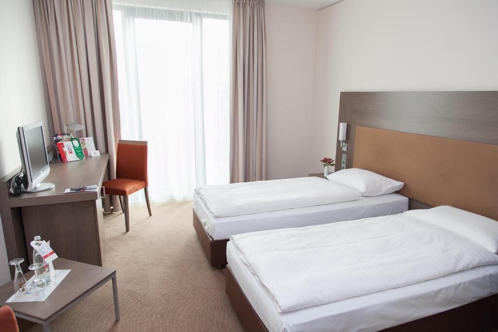 A hotel room with two beds, a desk with a computer, a chair, and a window with curtains.