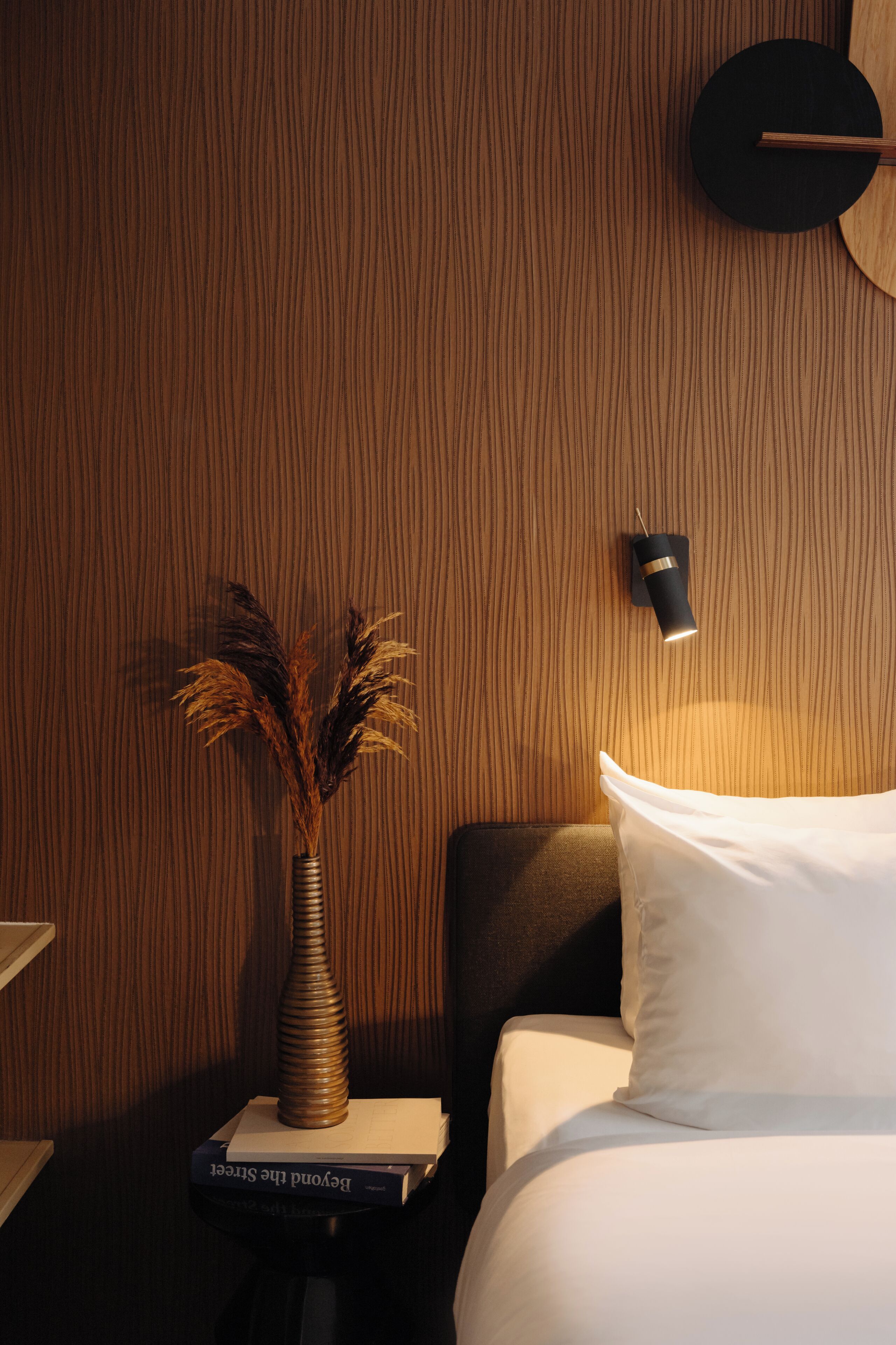 A neatly made bed with white linens, a bedside table with a vase of dried plants, and a wall-mounted clock.