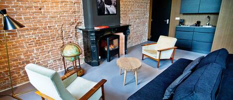 A living room with a brick wall, a fireplace, a globe, two chairs, a sofa, and a kitchen area with cabinets.