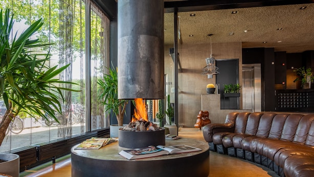 A modern lounge area with a circular seating arrangement, a central fire feature, and large windows providing natural light.