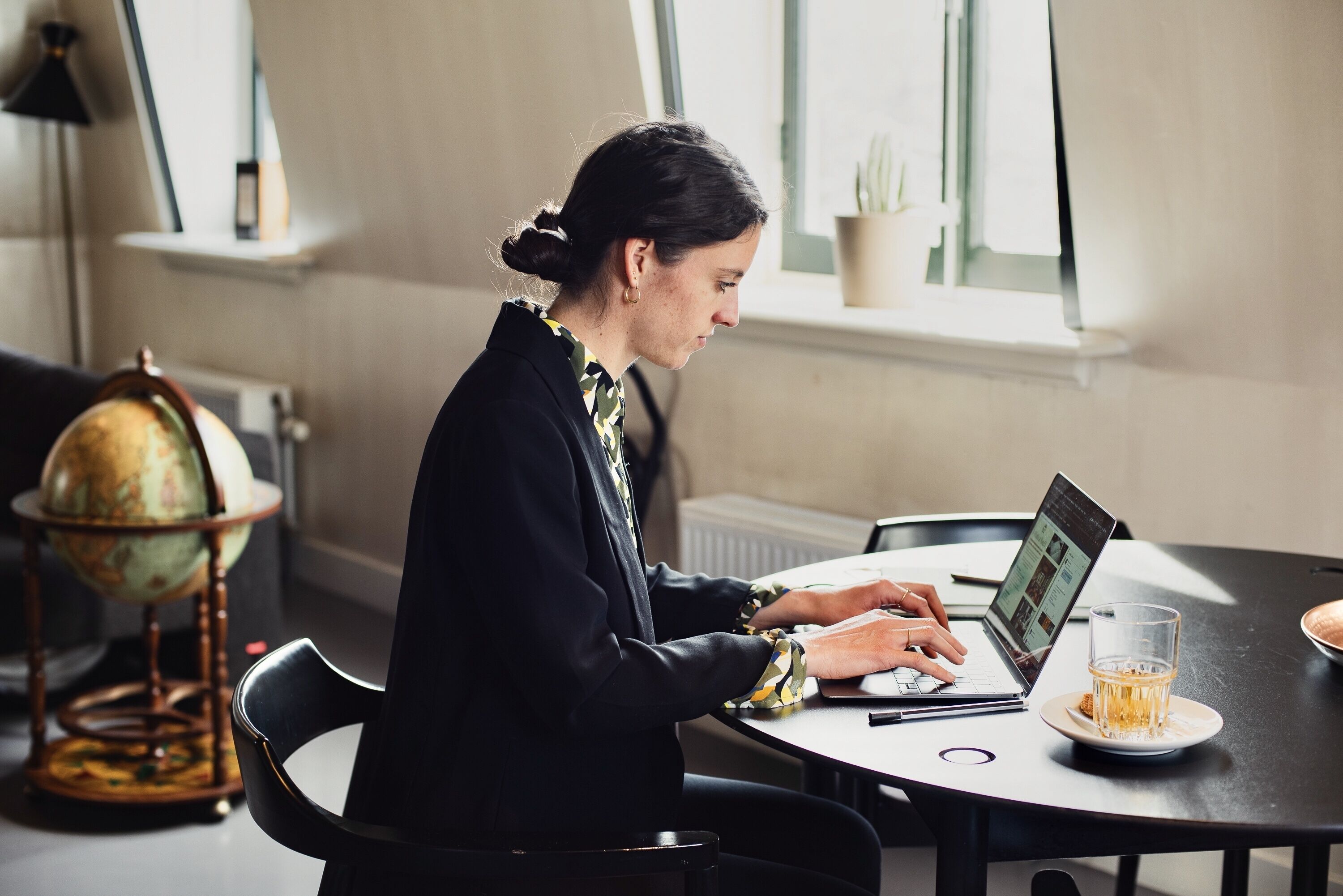 A person working at a desk with a laptop, a globe, and a glass of drink on the table.