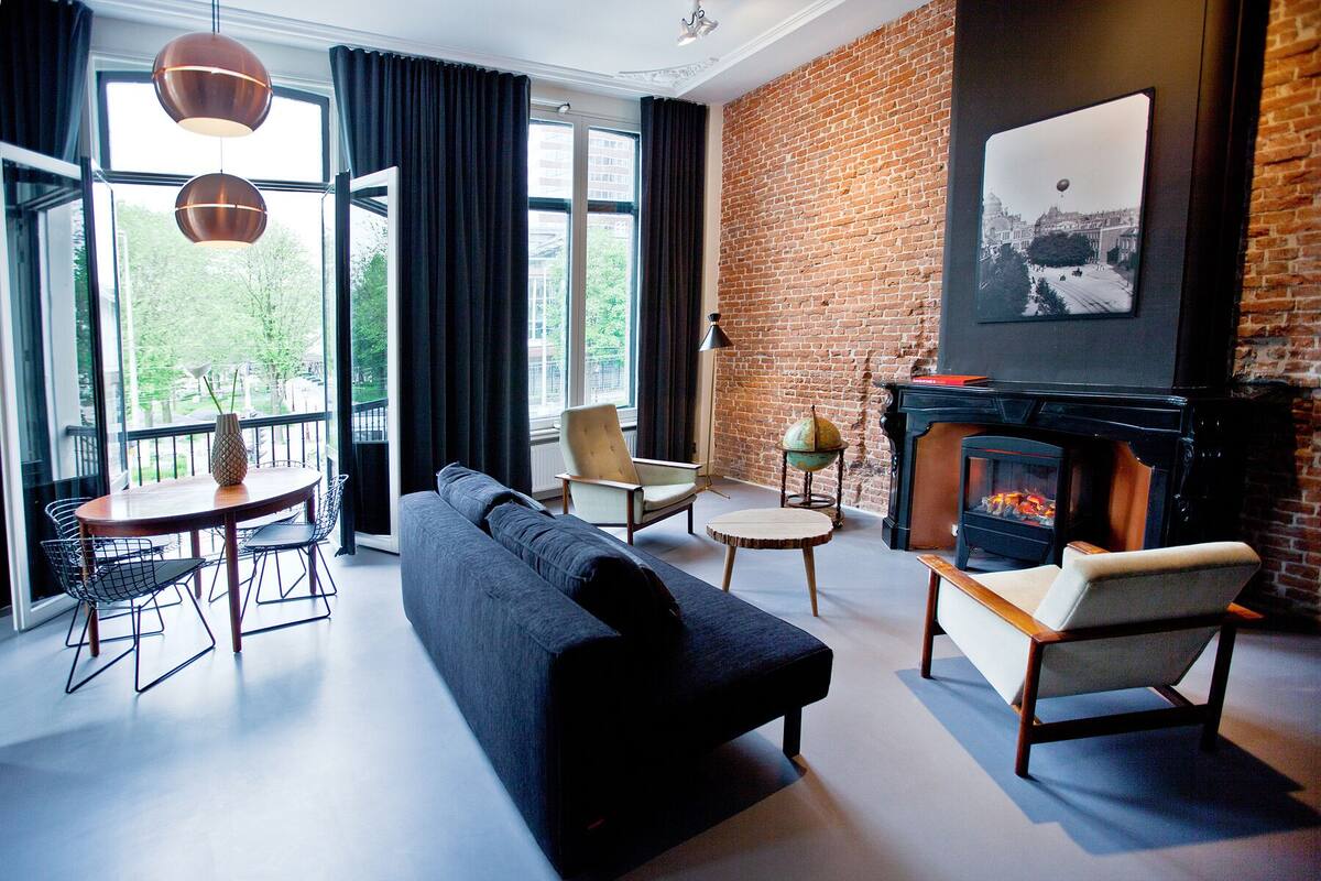 A modern living room with a black sofa, two armchairs, a round coffee table, and a fireplace. There is a large window with curtains, a globe on a stand, and a framed picture on the wall.