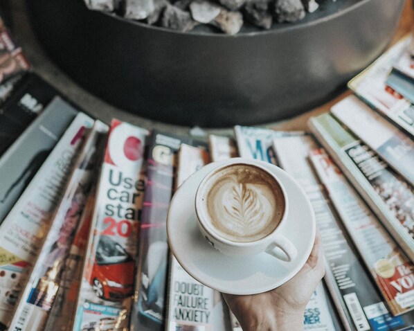 Une main tenant une tasse de café ornée d’un motif de latte art, entourée de magazines sur une table.