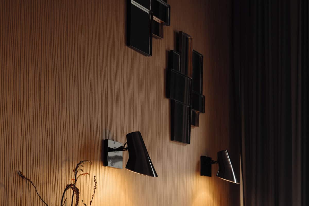 A hotel room with a bed, white pillows, a vase with plants, and wall-mounted lamps.