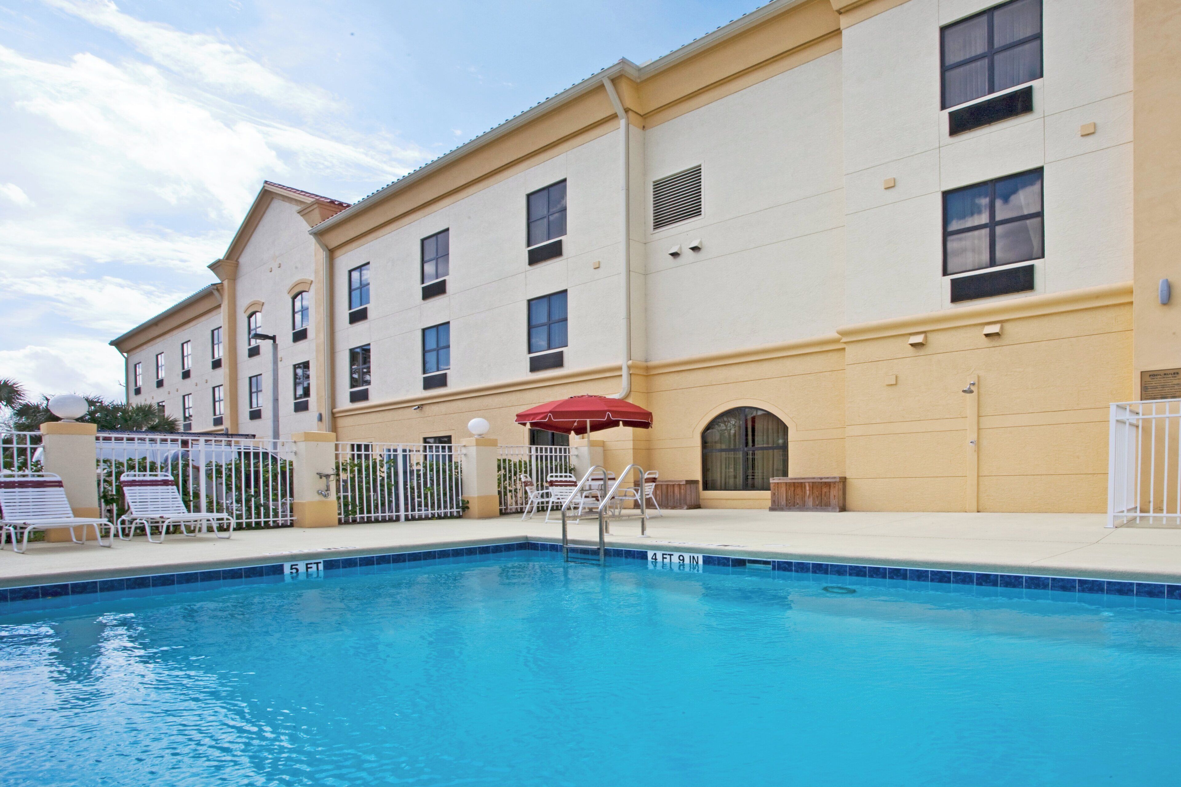A swimming pool with a red umbrella, white lounge chairs, and a building with multiple windows and balconies.