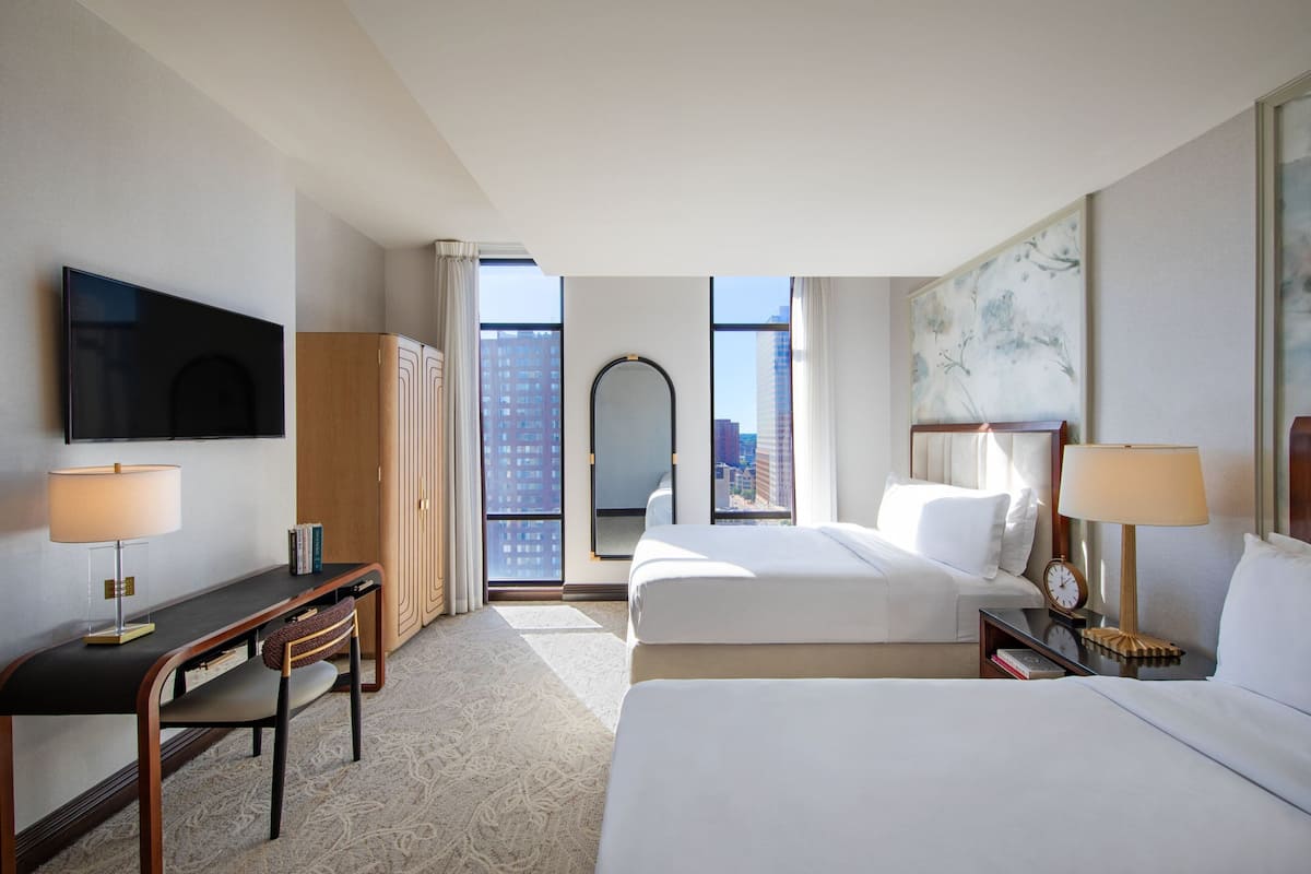 A hotel room with a bed, a desk, a chair, a television, a mirror, and a window with a city view.
